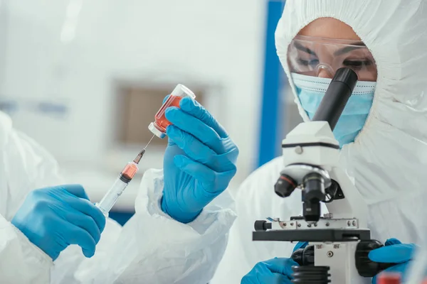 Vista recortada del bioquímico que toma medicamentos con jeringa cerca de su colega que trabaja con el microscopio - foto de stock
