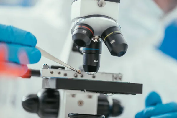 Vista parcial del bioquímico mirando sosteniendo un pequeño cálculo con pinzas cerca del microscopio - foto de stock