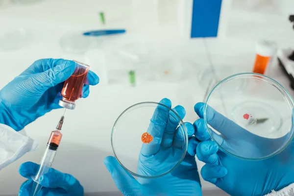 Vista recortada del bioquímico tomando medicamentos con jeringa cerca de su colega sosteniendo placas de Petri con biomaterial - foto de stock