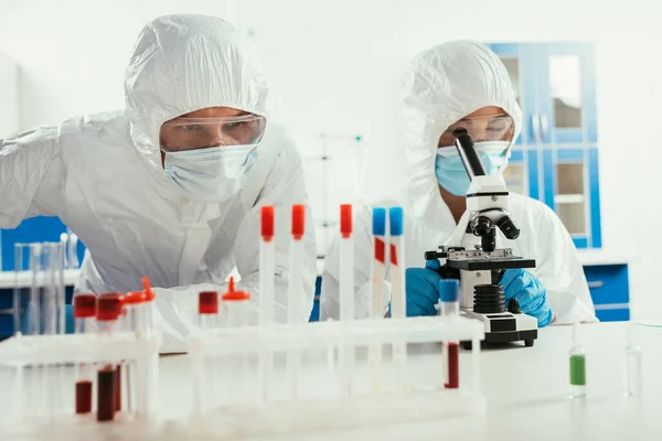 Bioquímico que trabaja con el microscopio cerca de colega que mira los tubos de ensayo en laboratorio - foto de stock