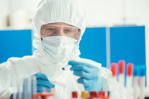 Bioquímico en traje de materiales peligrosos que sostiene la jeringa cerca de los tubos de ensayo en laboratorio - foto de stock