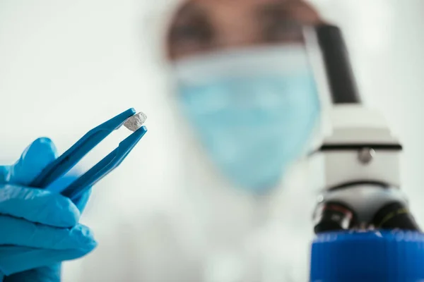 Foco seletivo do bioquímico segurando pequena pedra com pinças perto do microscópio — Fotografia de Stock