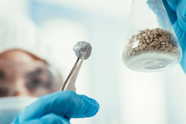 Foco seletivo do bioquímico segurando pequena pedra com pinça e frasco com cascalho — Fotografia de Stock