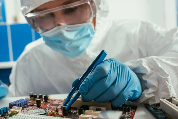Инженер в медицинской маске и очках, чинит материнскую плату компьютера пинцетом — стоковое фото
