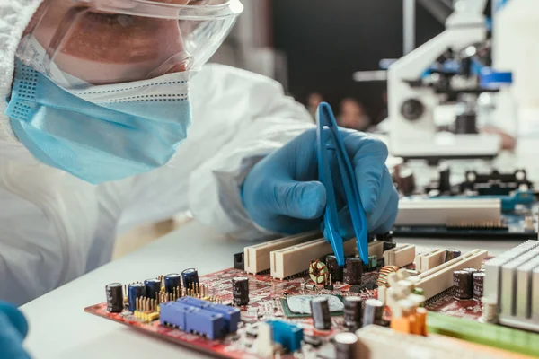 Engenheiro em máscara médica e óculos de fixação placa-mãe computador com pinças — Fotografia de Stock