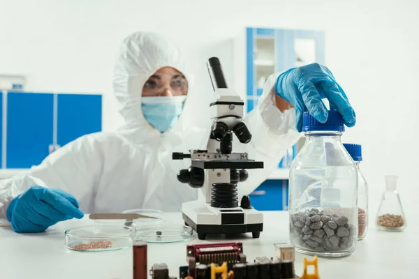 Engineer in hazmat suit holding jar with gravel near microscope and computer motherboard — Stock Photo