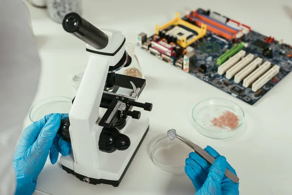 Partial view of engineer holding small stone with tweezers near microscope and computer motherboard — Stock Photo