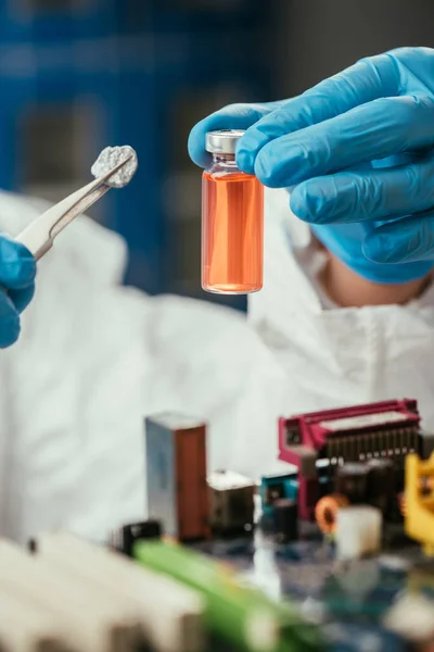 Vista ritagliata di ingegnere che tiene piccola pietra con pinzette e contenitore di vetro con liquido giallo vicino alla scheda madre del computer — Foto stock