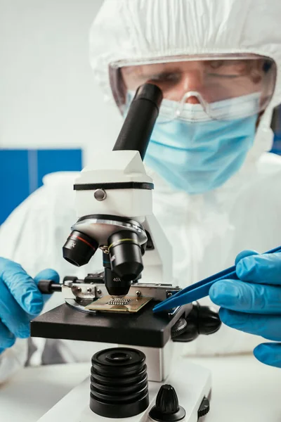 Engineer in hazmat suit looking at computer microchip through microscope — Stock Photo