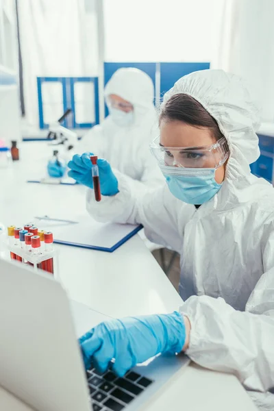 Biotecnólogo que sostiene el tubo de prueba con la muestra de sangre y usando el ordenador portátil - foto de stock