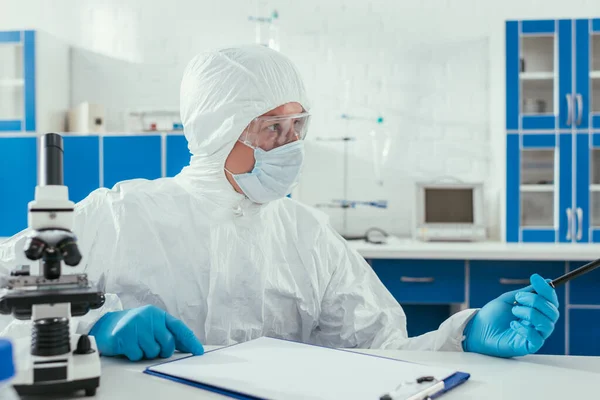 Bioquímico en traje de materiales peligrosos sentado cerca del microscopio y portapapeles en el laboratorio - foto de stock