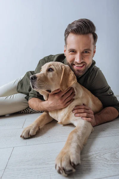 Enfoque selectivo del hombre sonriendo a la cámara mientras acaricia golden retriever en el suelo sobre fondo gris - foto de stock