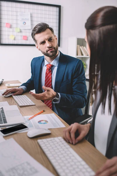 Selective focus of information security analyst looking at colleague near gadgets and papers with charts on table — Stock Photo