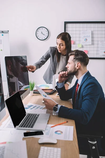 Analista de datos apuntando al monitor de computadora cerca de un colega sosteniendo papel con gráficos en la mesa - foto de stock