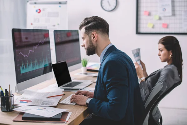 Selektiver Fokus des Datenanalysten, der mit Diagrammen am Computermonitor arbeitet, in der Nähe eines Kollegen mit Aufsatz im Büro — Stockfoto