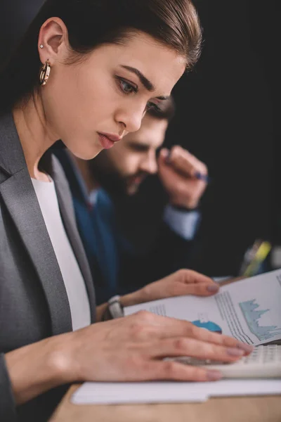 Focus selettivo dell'analista di dati che lavora con calcolatrice e grafici su carta vicino al collega su sfondo nero — Foto stock