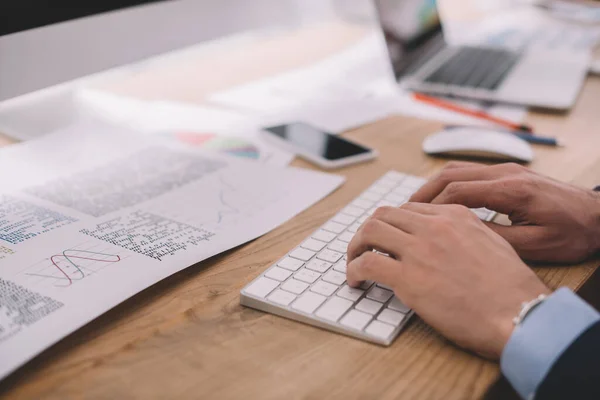 Vista recortada del analista de datos utilizando el teclado de la computadora cerca de papeles con gráficos en la tabla - foto de stock