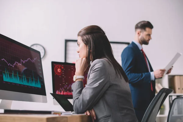 Selective focus of data analyst looking at graphs on computer monitor near colleague with paper in office — Stock Photo