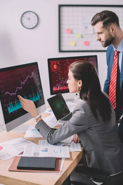Side view of data analyst pointing with hand at graphs on computer monitor to colleague in office — Stock Photo