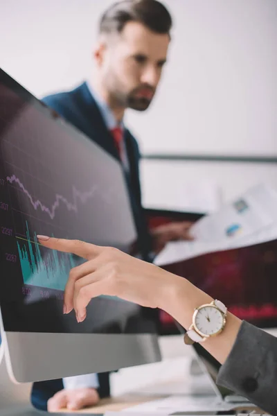 Enfoque selectivo del analista de datos apuntando a gráficos en monitor de computadora cerca de colega con papeles en la oficina — Stock Photo