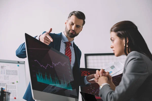 Selective focus of data analyst with papers pointing on graphs on computer monitor to colleague in office — Stock Photo