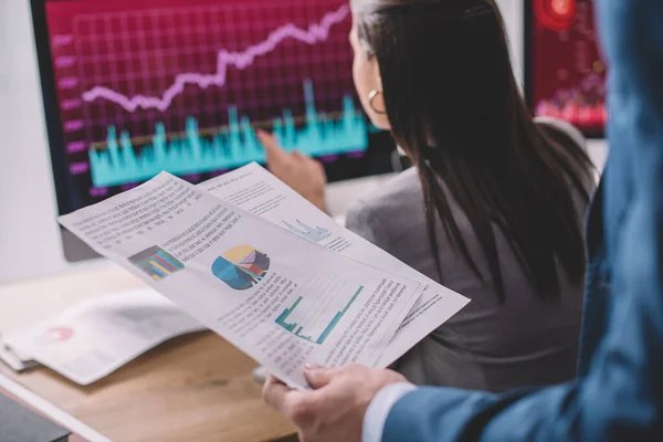 Enfoque selectivo del analista de datos que sostiene papeles con gráficos cerca de un colega que trabaja con computadoras en una mesa aislada en gris — Stock Photo