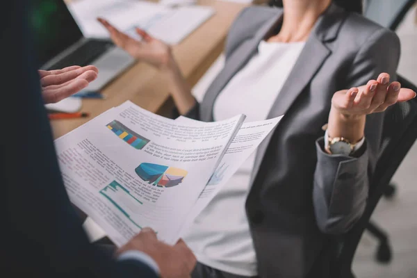 Cropped view of data analyst holding papers with graphs near colleague showing dubium gesture at table — Stock Photo