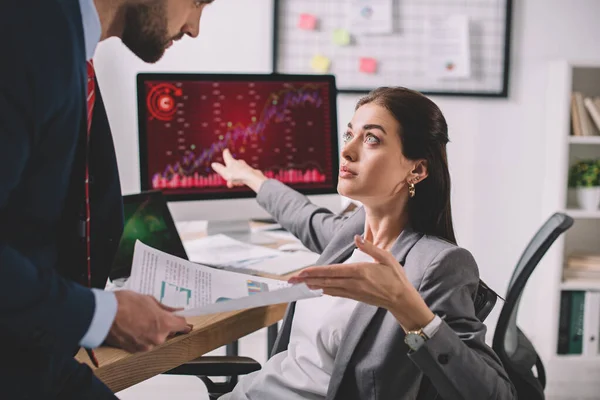 Side view of data analyst pointing on computer monitor with charts near colleague with papers — Stock Photo