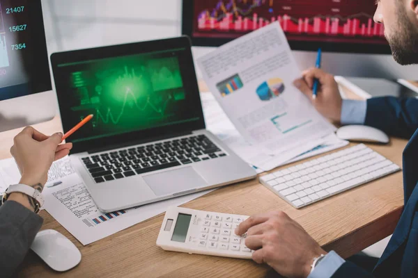 Cropped view of information security analysts using calculator and graphs on computer monitors while working in office — Stock Photo