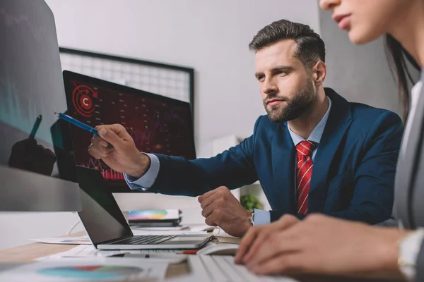 Selective focus of data analyst pointing on graphs on computer monitor to colleague at table — Stock Photo