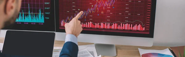 Cropped view of data analyst pointing on charts on computer monitor near laptop with blank screen on table, panoramic shot — Stock Photo