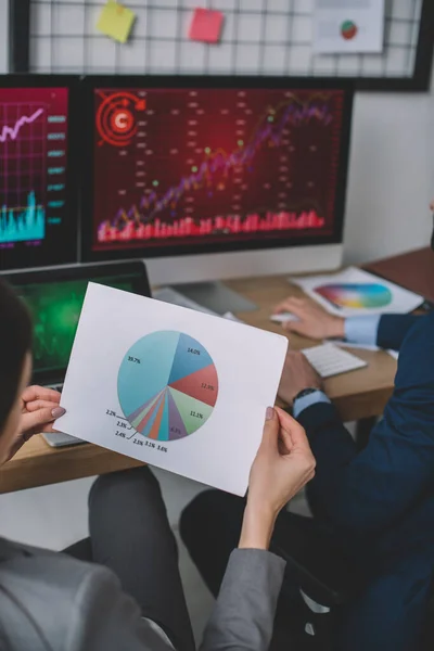 Vista recortada del analista de seguridad de la información sosteniendo papel con gráfico cerca de colega utilizando la computadora en la tabla - foto de stock