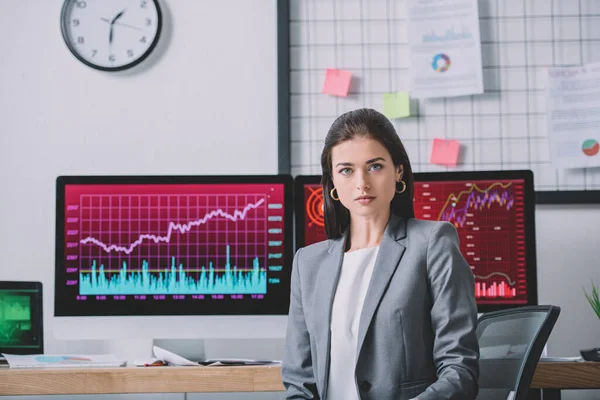 Beautiful data analyst looking at camera near charts on computer monitors in office — Stock Photo