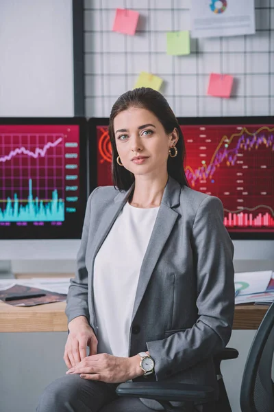 Beautiful information security analyst looking at camera near graphs on computer monitors on table — Stock Photo