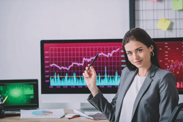 Analyste de données regardant la caméra et pointant vers les graphiques sur les écrans d'ordinateur dans le bureau — Photo de stock