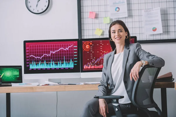 Smiling computer systems analyst looking at camera near charts on computer monitors on table — Stock Photo