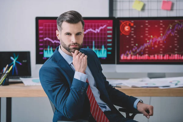 Analista de datos guapo mirando la cámara cerca de gráficos en monitores de computadora en la mesa en la oficina - foto de stock