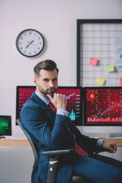 Analista de datos guapo mirando la cámara cerca de gráficos en monitores de computadora en la oficina - foto de stock