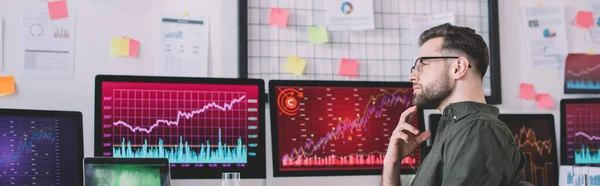Side view of pensive data analyst looking away near charts on computer monitors in office, panoramic shot — Stock Photo