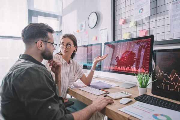 Data analyst pointing with hand at charts on computer monitor to colleague at table in office — Stock Photo