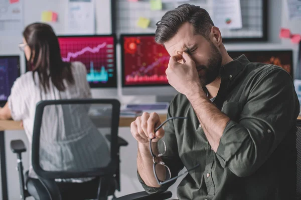 Selective focus of tired data analyst holding eyeglasses near colleague working with computers in office — Stock Photo