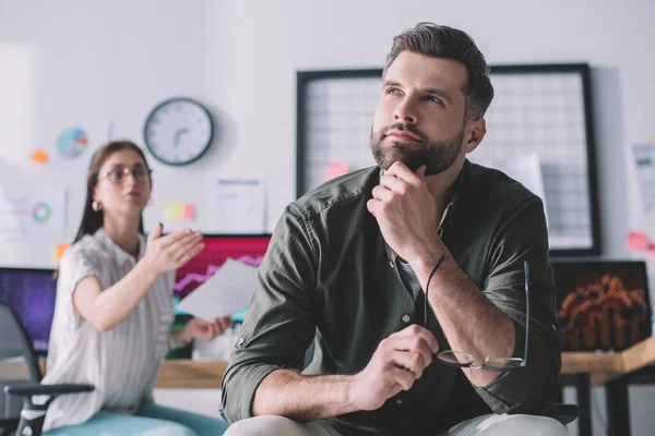 Selective focus of dreamy data analyst holding eyeglasses near colleague in office — Stock Photo