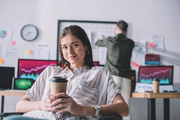 Analyste de données souriant tenant un café à emporter pendant qu'un collègue travaille au bureau — Photo de stock