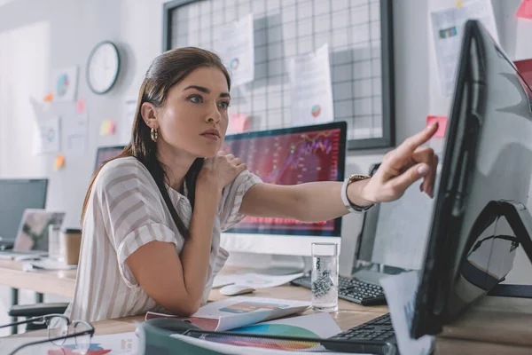 Selective focus of data analyst pointing with finger at computer monitor near papers and glass of water on table — Stock Photo