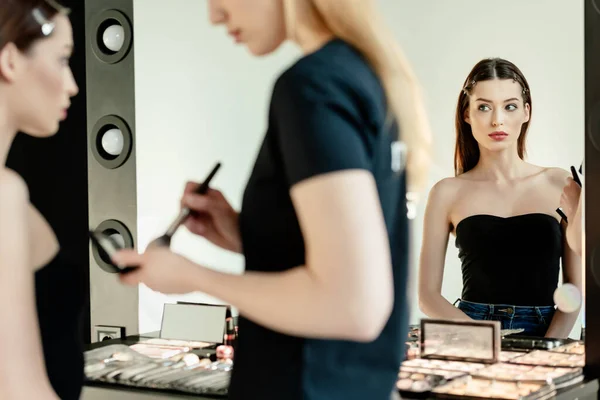 Selective focus of beautiful model looking at mirror near makeup artist — Stock Photo