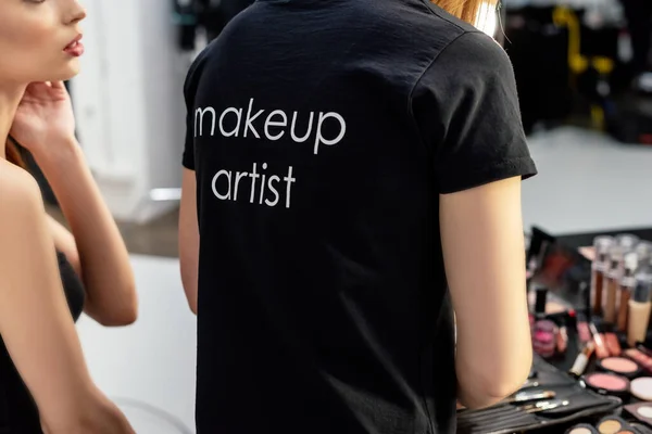 Ausgeschnittene Ansicht einer Frau im schwarzen T-Shirt mit dem Schriftzug des Make-up Artist in der Nähe des Modells — Stockfoto