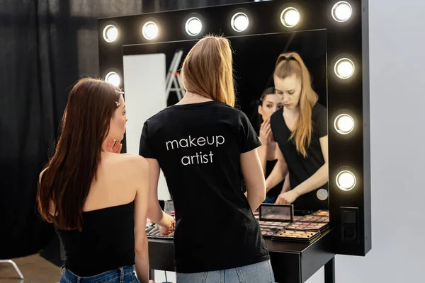 Mulher em t-shirt preta com maquiagem artista lettering pé perto de modelo e cosméticos decorativos — Fotografia de Stock