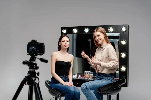 Selective focus of happy makeup artist holding lip gloss near model and digital camera isolated on grey — Stock Photo