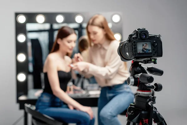 Enfoque selectivo de la cámara digital cerca de maquillaje artista celebración de brillo de labios cerca de modelo aislado en gris - foto de stock