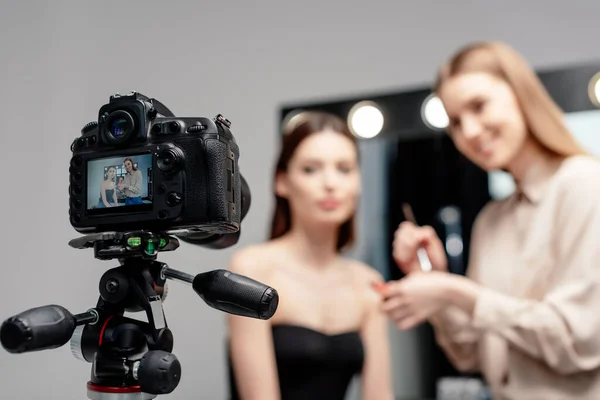 Foco seletivo da câmera digital com artista de maquiagem alegre segurando brilho labial perto do modelo na tela isolada em cinza — Fotografia de Stock